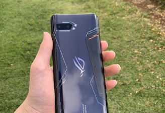 A hand holding a sleek, dark-colored smartphone with geometric patterns and the text 'Tencent Games' on the back cover. The background features a grassy area with some trees or bushes on the side.